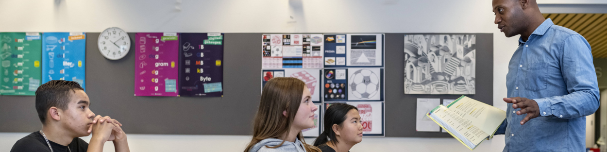 Leerlingen zitten in de klas en krijgen les. Leraar geeft uitleg aan de leerlingen.